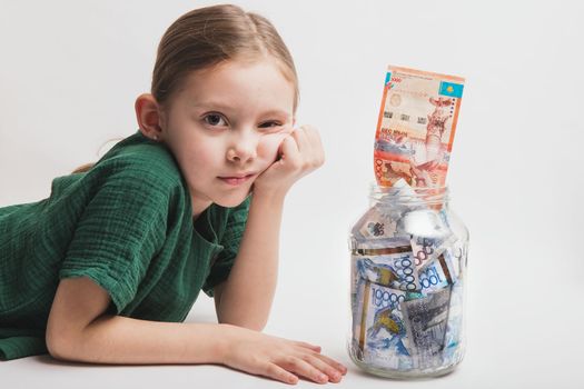 Sad and bored child with a large transparent jar with Kazakhstani tenge money on a white background.
