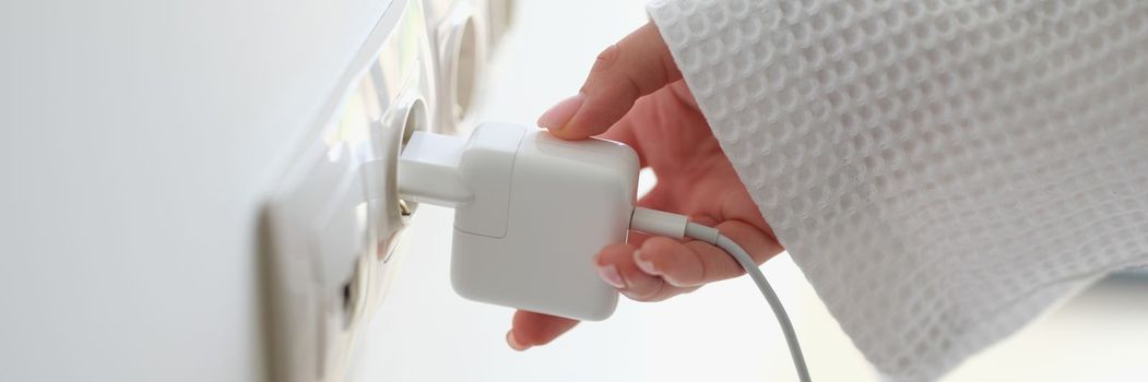 Woman hand in robe gown inserts white charger into socket in hotel rooms or at home. Charge gadgets correctly concept
