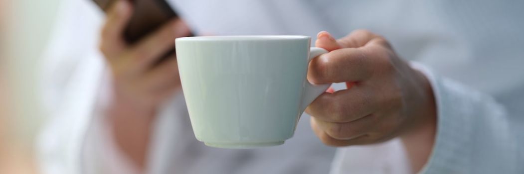 Cropped photo of a woman in a terry bathrobe holding mobile phone and cup. Watching news and relaxing morning coffee