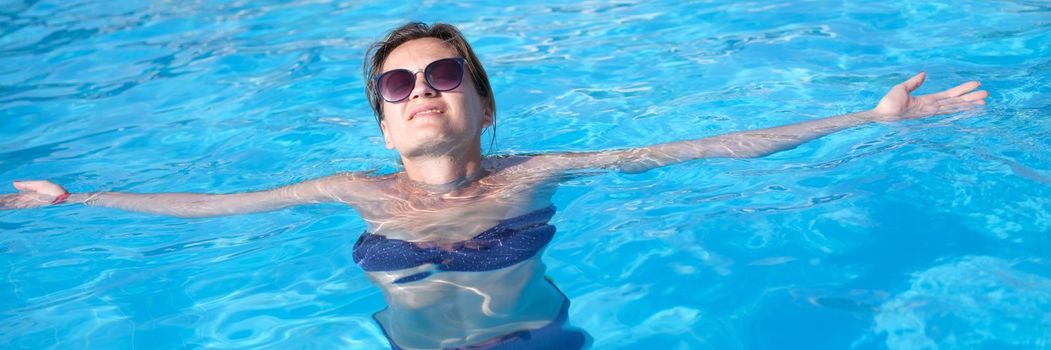 Charming woman in sunglasses enjoys swimming in turquoise water of pool. Relax in luxury hotel pool and wellness concept