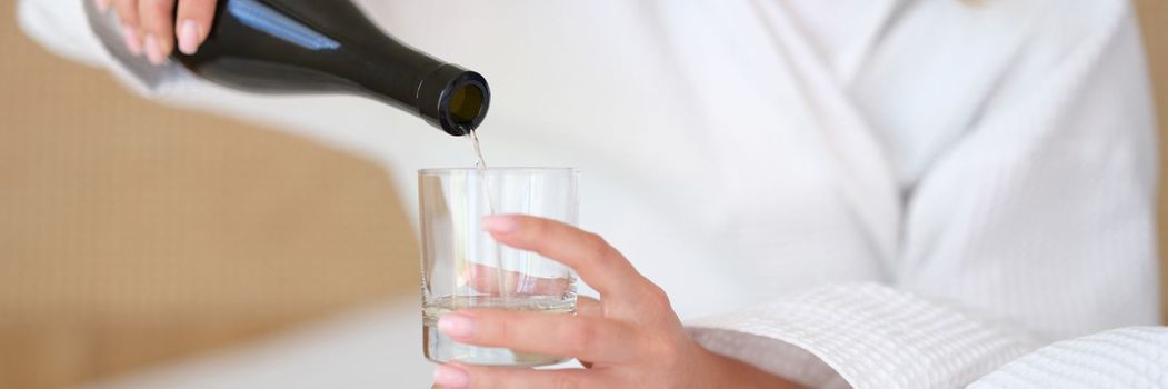 Woman in bathrobe on bed pours water or white wine into glass alone. Glass of water in morning on empty stomach and alcoholism alone concept