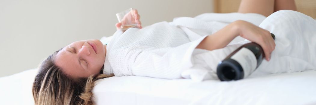 Drunk woman in bathrobe sleeps on bed with bottle of wine and glass in hotel room. Female alcoholism stress life problems or drunk female tourist on vacation