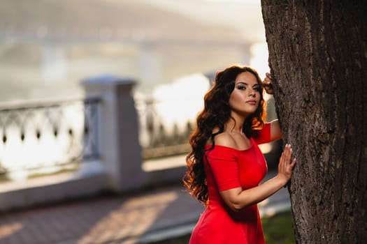 Close-up portrait of a beautiful young girl with long dark wavy hair in a red dress at the background of summer sunset