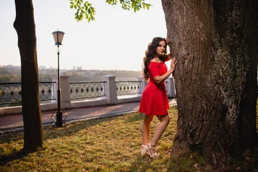 Portrait of a beautiful young girl with long dark wavy hair in a red dress at the background of summer sunset
