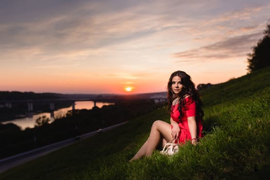Beautiful young girl with long dark wavy hair sitting on a slope covered with green grass against the background of a summer sunset