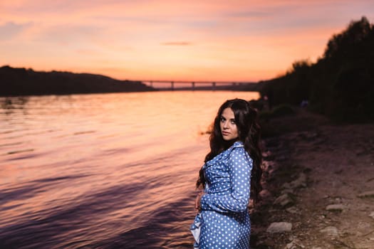 Beautiful young girl with long dark wavy hair standing at the bank of the river against the background of a bright sunset