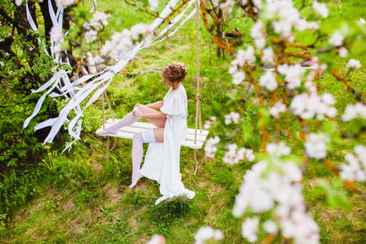 Young bride with blond hair in white negligee and stockings sitting on a rope swing putting on a garter at the background of spring blossom orchard