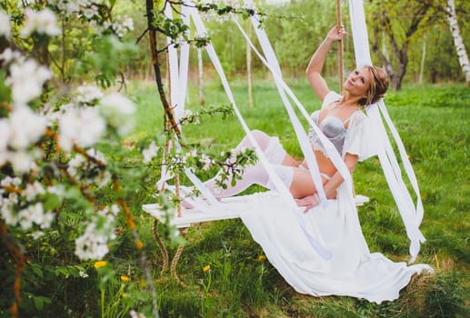Young bride with blond hair in white negligee and stockings sitting on a rope swing at the background of spring blossom orchard