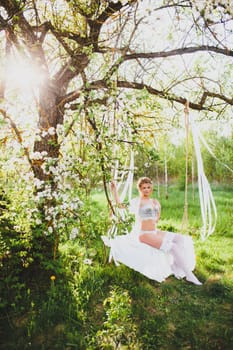 Young bride with blond hair in white negligee and stockings posing on a rope swing at the background of spring blossom orchard