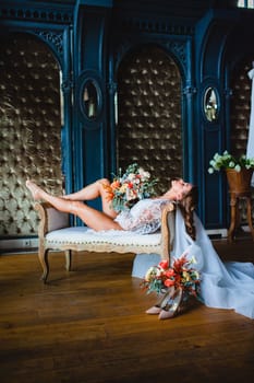 Seductive young bride with beautiful naked legs in white negligee and lingerie posing with gorgeous bridal bouquet on a sofa