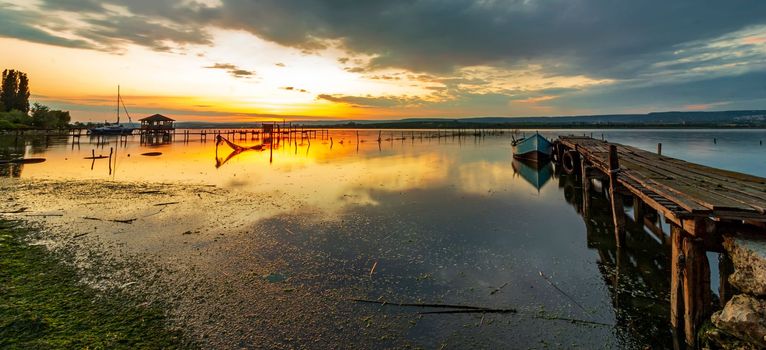 sunset at a lake coast with a boat at a wooden pier. Panoramic view