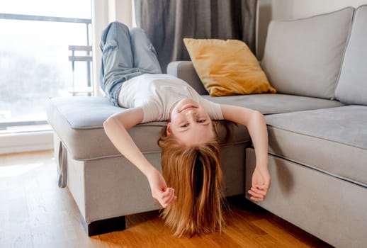 Preteen girl lying on sofa with head down and posing looking at camera at home. Pretty child kid on couch in living room