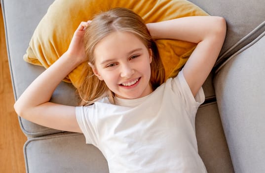 Preteen girl lying on sofa cushion and posing looking at camera at home. Pretty child kid on couch in living room portrait
