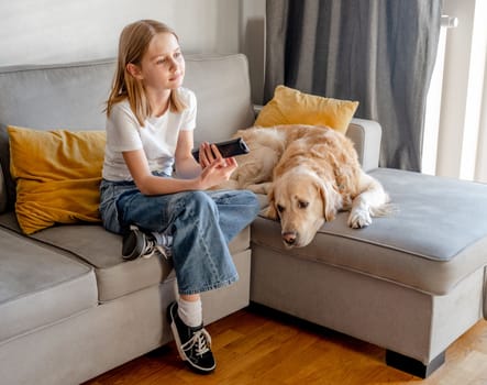 Preteen girl with golden retriever dog holding tv remote control and sitting on sofa at home. Beautiful child with labrador doggy pet watching cartoons at television kids channel