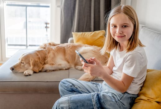 Preteen girl with golden retriever dog holding smartphone and sitting on sofa at home. Beautiful child kid with labrador doggy pet using cell phone for social media connection