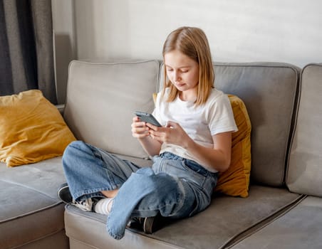 Preteen girl with smartphone chatting with friends on sofa at home. Pretty child kid using modern mobile cellphone for communication