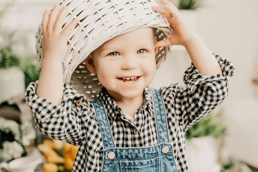 Little girl 1 years old with a basket on her head. Funny child plays puts a basket on her head like a hat and laughs, she is dressed in a plaid shirt and denim overalls. Funny moments with kids