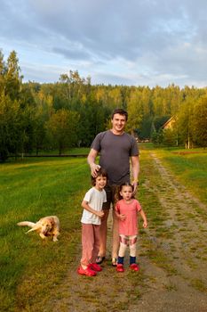 Adult man with daughter and son standing on path near loyal dog on summer weekend day in nature