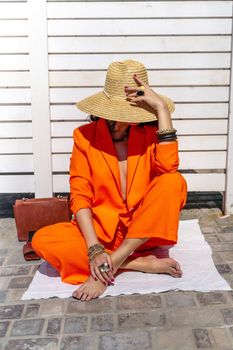 Stylish woman in an orange suit with a hat sits on a rug on a white striped background. On the hands are jewelry rings and bracelets, sandals and a bag stand side by side