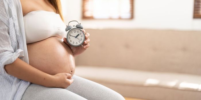 An Asian pregnant woman in comfortable clothes sits on a chair in her living room, holding her belly and alarm clock. awaiting baby, baby's due date concept.