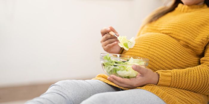 Happy pregnant young woman sitting and eating salad at home.