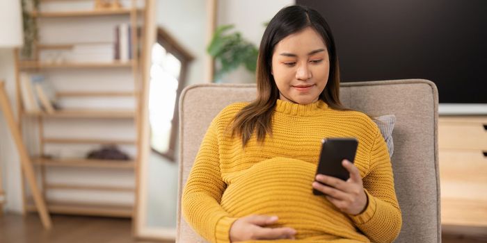 Happy Asian pregnant woman using her phone while relaxing on sofa in her living room.