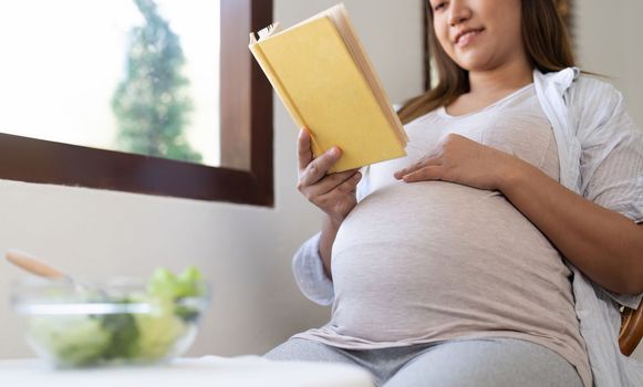 Young pregnant woman sitting reading a book and eating salad at home.