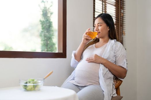 Happy pregnant young woman sitting and eating salad and orange juice at home.