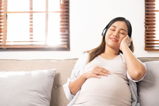 Happy Asian pregnant woman listening to music through her headphones on sofa. pregnancy lifestyle.