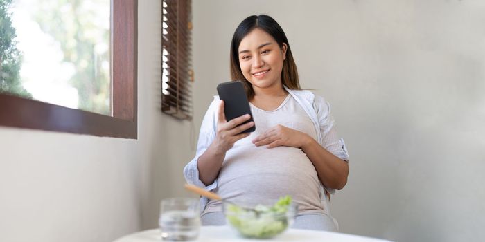 Young pregnant woman sitting using phoneand eating salad at home.