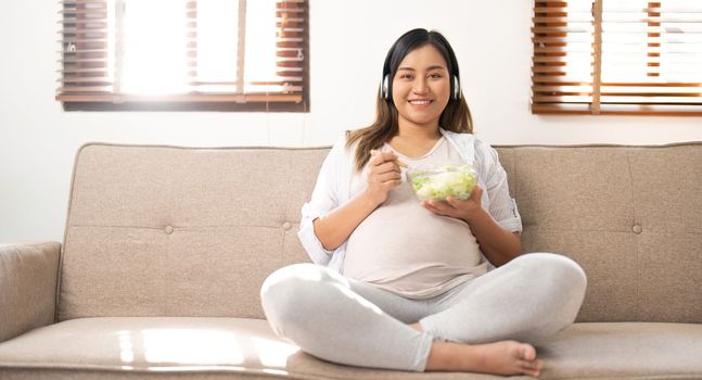 Relaxed Asian pregnant woman eating a healthy salad bowl on sofa in the living room.