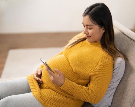 Happy Asian pregnant woman using her phone while relaxing on sofa in her living room.