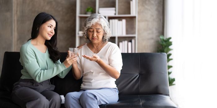 Asian daughter giving pills to mother helping her mom taking medicine with glass of water.