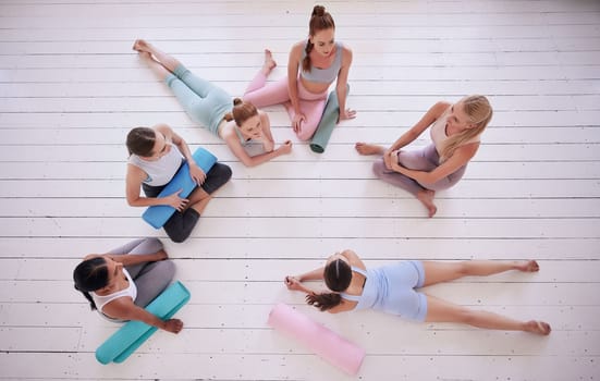 Group of women relaxing in yoga class from above. Friends talking, being social in pilates class. Women bonding in yoga class. Young women sitting on the floor in yoga class from above.