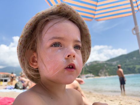 Little girl with sunscreen on her face sits on the beach under sun umbrella. High quality photo