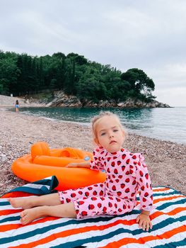 Little girl sits on a blanket on a pebble beach with her hand on an inflatable ring. High quality photo