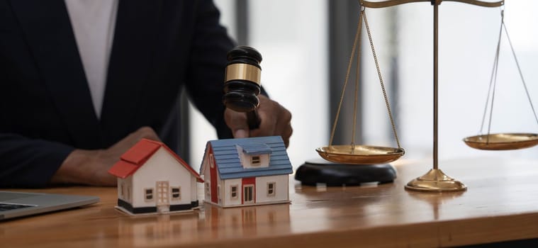 Close-up of a house in front of a lawyer holding a hammer and a laptop silver brass scales on a wooden table in his office, law, legal services, advice, justice and real estate ideas...