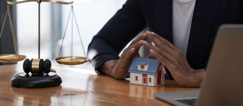 Close-up of a house in front of a lawyer holding a hammer and a laptop silver brass scales on a wooden table in his office, law, legal services, advice, justice and real estate ideas...