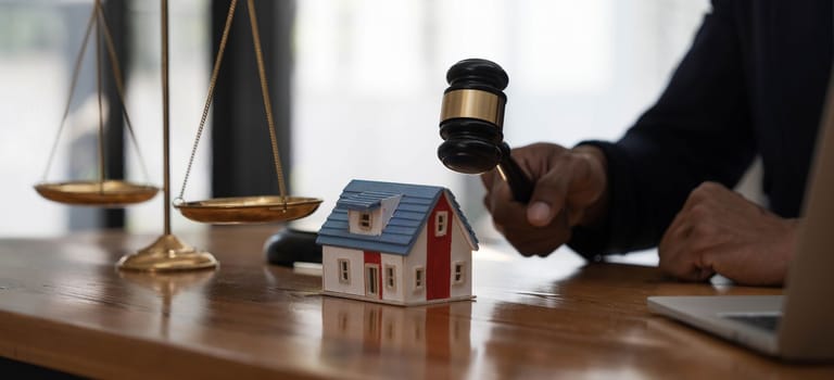 Close-up of a house in front of a lawyer holding a hammer and a laptop silver brass scales on a wooden table in his office, law, legal services, advice, justice and real estate ideas...