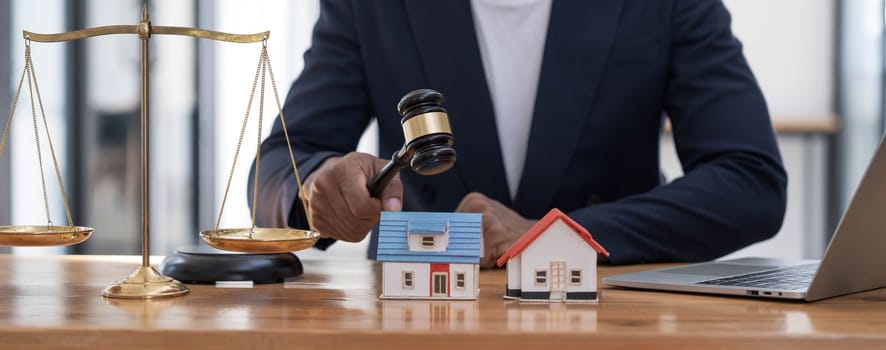 Close-up of a house in front of a lawyer holding a hammer and a laptop silver brass scales on a wooden table in his office, law, legal services, advice, justice and real estate ideas...