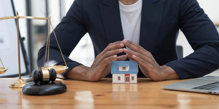 Close-up of a house in front of a lawyer holding a hammer and a laptop silver brass scales on a wooden table in his office, law, legal services, advice, justice and real estate ideas...