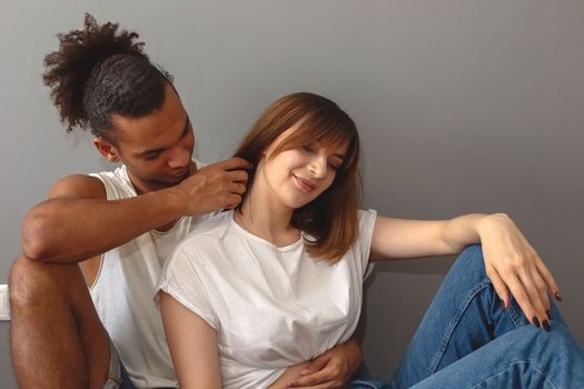 Multiracial couple , guy and girl in white t-shirts sit at home on the floor against a gray wall, gentle touches.