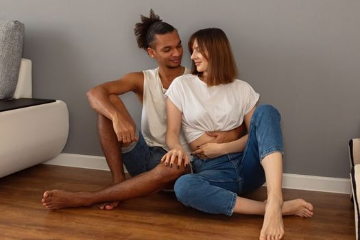 A stylish multiracial couple, a guy and a girl in white t-shirts and blue jeans, sit on the floor at home against a gray wall, hugging. Copy space