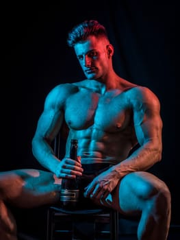 Muscular naked shirtless sexy young man with champagne bottle, sitting on floor, isolated on black background