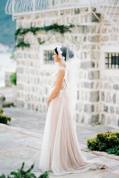 Bride in a veil stands near an old stone building. Side view. High quality photo
