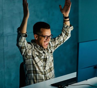 Portrait of a young happy businessman by monitor and desktop computer celebrating success and victory working late at night in office