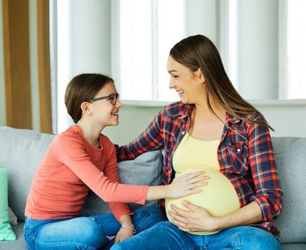 Portrait of a happy young pregnant mother and her daughter holding her belly at home