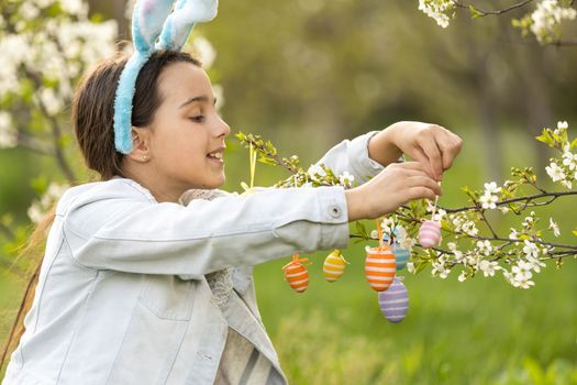 Adorable little girl in bunny ears, blooming tree branch outdoors on a spring day. Kid having fun on Easter egg hunt in the garden.