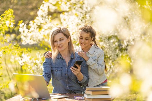 Happy girl and mom relaxing with gadgets outdoor. Mother and little daughter kid using parental control app on smartphone, playing online game, watching video on laptop, browsing internet.