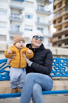 Young mother with her cute infant baby boy child on bench in city park